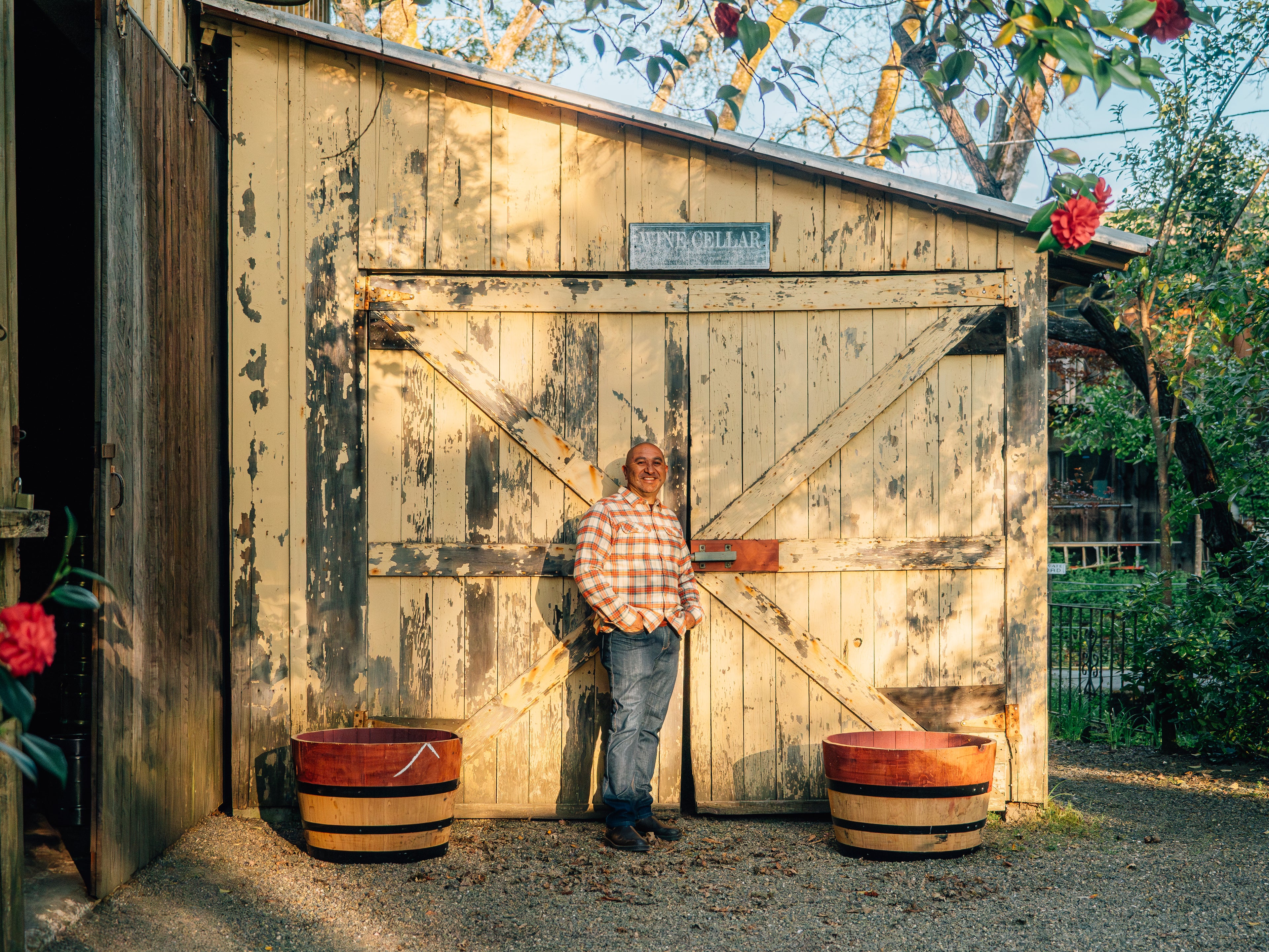 Man standing in front of a wooden shed with two barrels, surrounded by flowers.